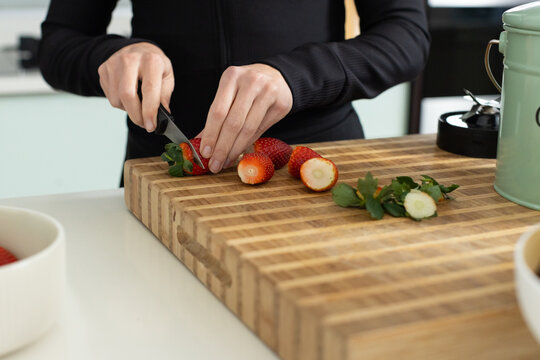 Woman slicing strawberries on butcher block on counter using paring knife in black top in kitchen
