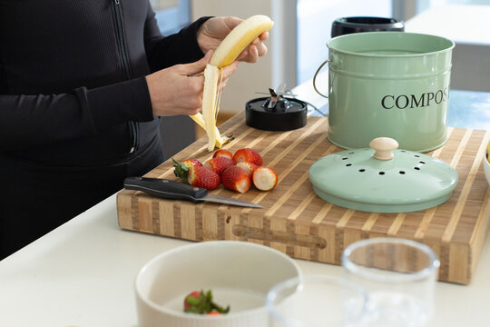 Woman wearing black zip-up top standing at kitchen counter peeling banana over butcher-block board