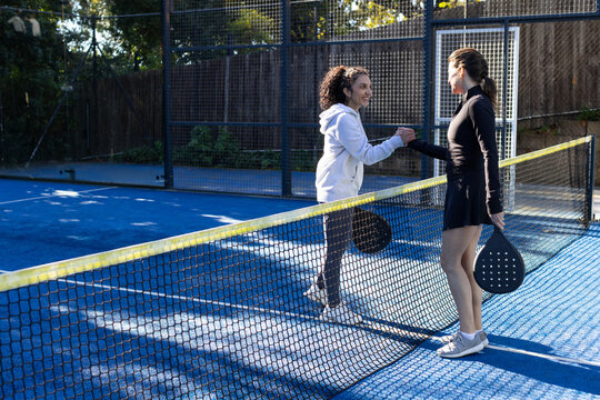 Diverse female players meeting at padel net on blue court, exchanging fist bump with rackets