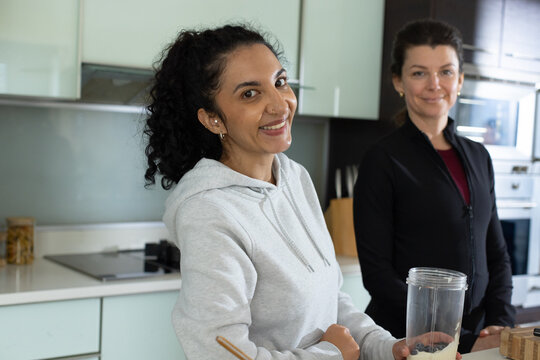 Diverse female friends wearing hoodie and zip jacket stirring smoothie in kitchen, with blender cup