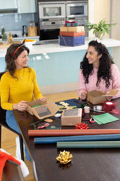 Diverse female friends wearing antler bands wrapping gifts at kitchen table with kraft boxes