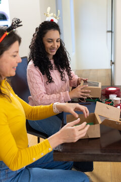 Female friends wrapping kraft boxes with clear lids and ribbons at table wearing antler headbands