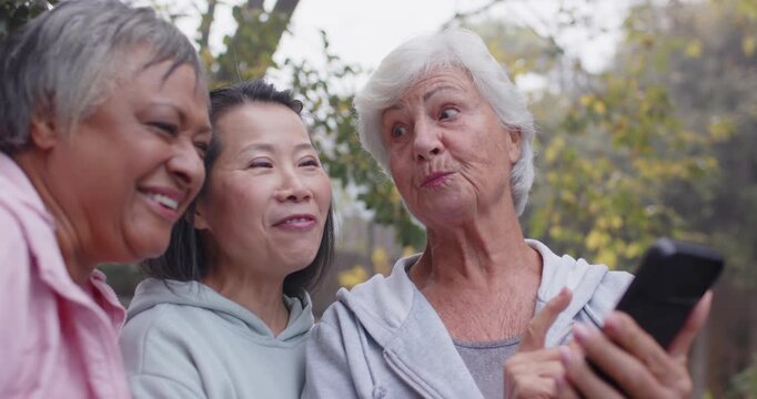 Diverse women friends leaning senior woman angling phone tapping causing laughs with bottle in park