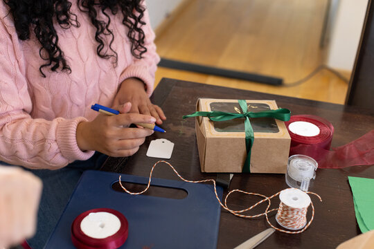 African American woman wearing pink sweater writing gift tag with blue pen beside kraft box