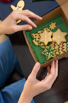 Woman arranging holiday sugar cookies into cardboard box with green napkin on home table in jeans