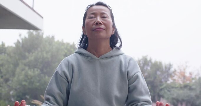 Mature Asian woman settling on yoga mat on deck in hoodie, lifting chin, closing eyes, meditating