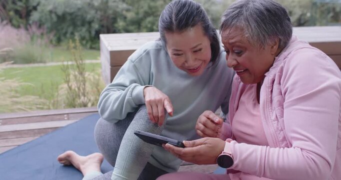 Asian, African American women in activewear leaning toward tablet tapping app for workout on deck