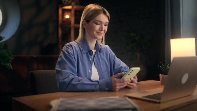 Cheerful young blonde woman scrolling social media feed on smartphone and laughing, happy female freelancer checking funny memes or positive news on phone at desk with laptop in cozy room.