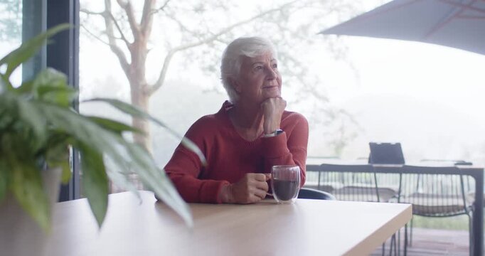 Senior woman in red sweater holding clear glass mug at table, noticing mist outside and reflecting