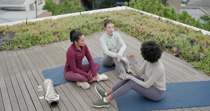 Diverse female friends showing phone, laughing and leaning on blue mats on roof deck post-workout