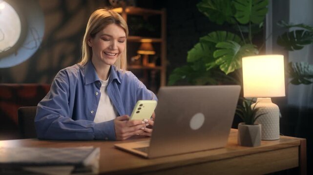Happy young blonde woman laughing and sharing funny memes on smartphone with friends, cheerful female freelancer texting and sending messages on phone at desk with laptop in cozy room.
