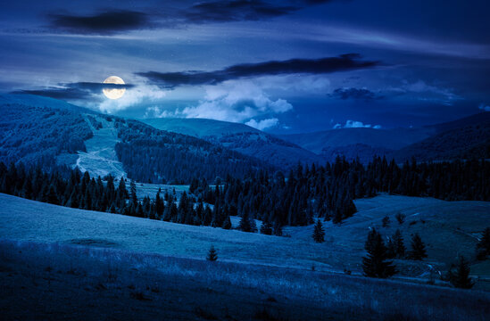 mountainous countryside landscape on summer night. full moon over view of green rural scenery with grassy pastures and rolling forested hills. mysterious mountain weather with cloudy sky