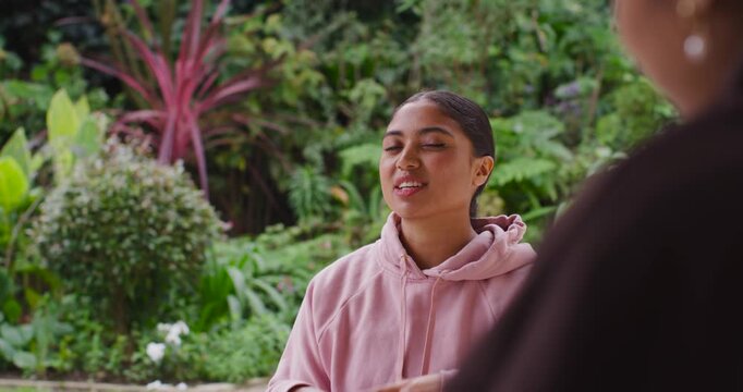 Diverse female pair with speaker in pink hoodie responding to prompt, gesturing on garden patio