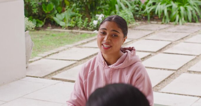 Indian woman in pink hoodie being prompted by friend, speaking and shifting moods on porch bench