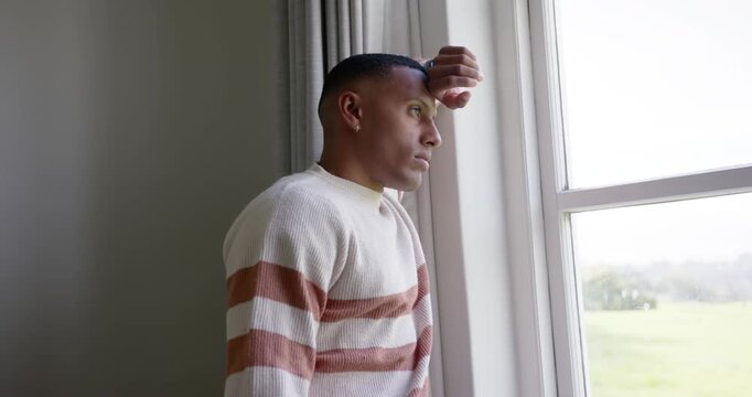 African American man standing by window in striped sweater raising right hand to brow reflecting