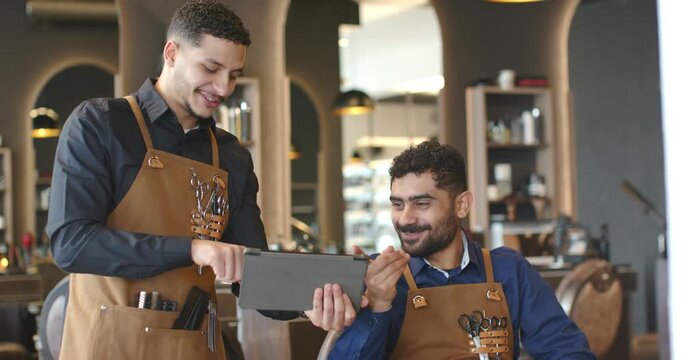 African American male barbers in aprons presenting tablet at salon station, guiding styling options