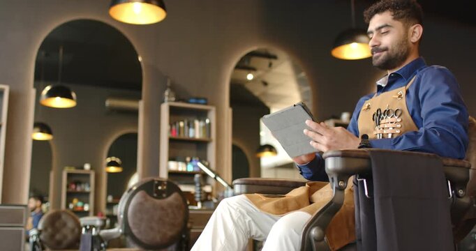 Male barber in shop apron using tablet, scrolling, glancing smiling as camera moving, copy space