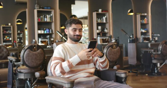 Man sitting in barber chair at salon leaning back, checking smartphone while waiting for haircut