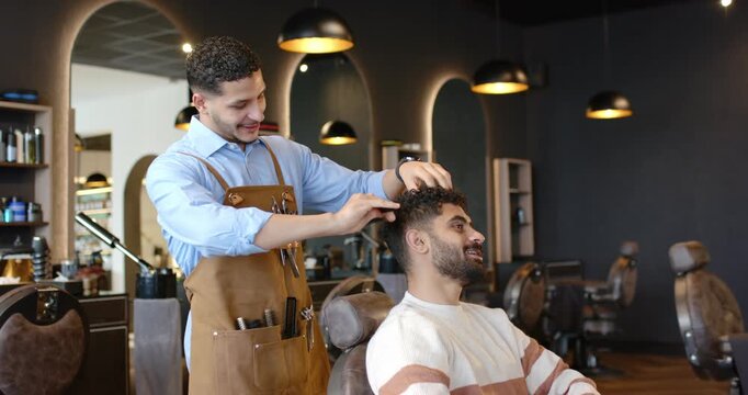 African American barber in leather apron cutting seated client with comb, scissors in barbershop