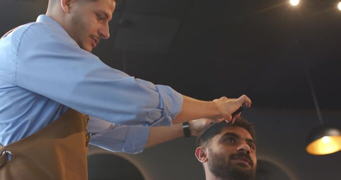 Male barber trimming male client's hair with trimmer and comb in barbershop after client request