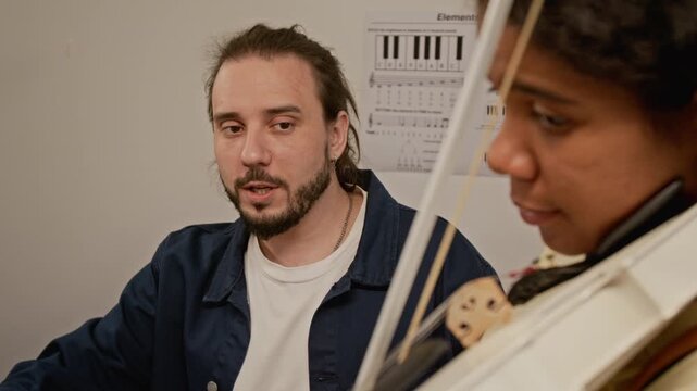 Rack focus shot of Caucasian young male teacher instructing Black teenager playing electric violin. Video for music school advertising and online learning platforms