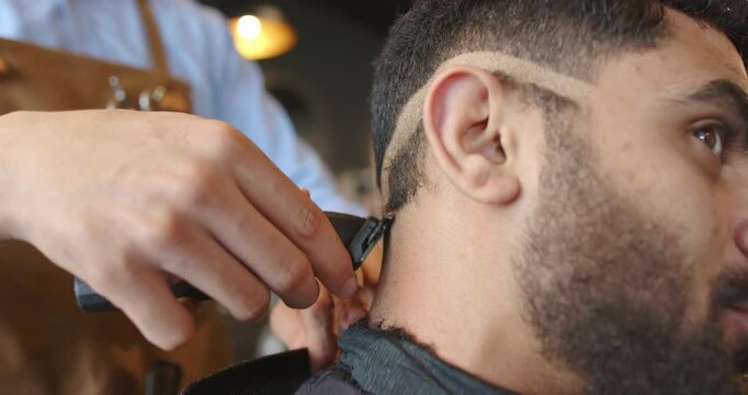 Barber in leather apron positioning clipper at man's neck at shop, trimming, refining fade