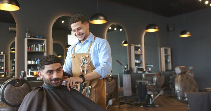 Barber and client, male in apron trimming sideburns with clippers in shop after client turning head
