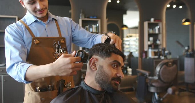 Male barber and client, in apron cutting with clipper as client tilting creating fade at shop