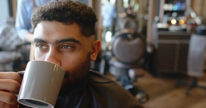 African American male client sipping gray mug while male barber moving in, starting clipper cut