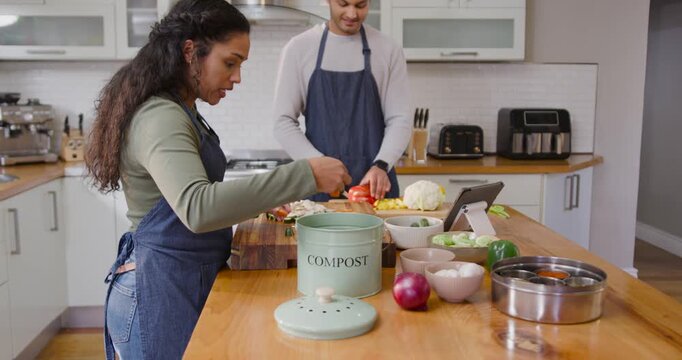 African American couple using tablet prepping veg on island using COMPOST for meal in aprons