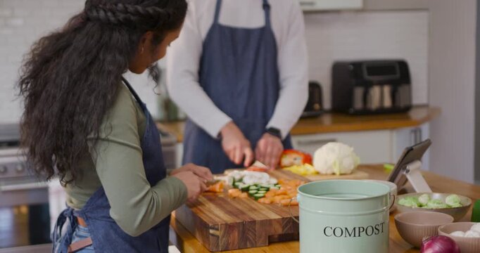 African American couple in aprons chopping peppers at island using tablet preparing meal, compost
