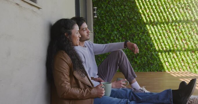 Diverse couple sitting on wooden deck, holding ceramic mugs while sunlight slats shifting, relaxing