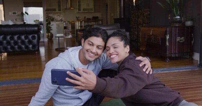 African American couple sitting on hardwood floor, man holding blue phone outward, reviewing selfie
