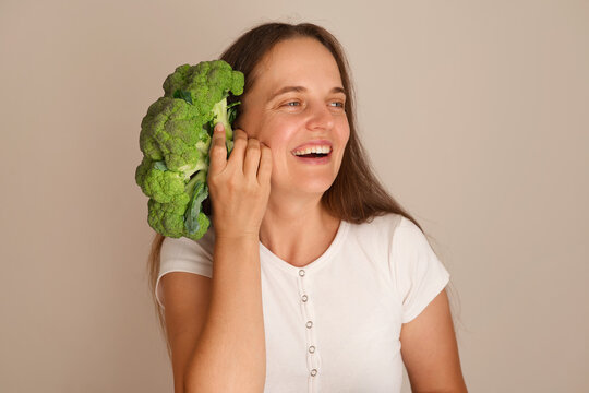 A woman stands with broccoli close to her ear and smiles. She wears a simple shirt and poses against a plain background. Her long hair is loose
