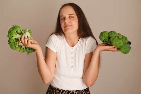 A woman stands with a piece of broccoli in each hand. She has a light expression while looking at the camera. The background is plain and light-colored