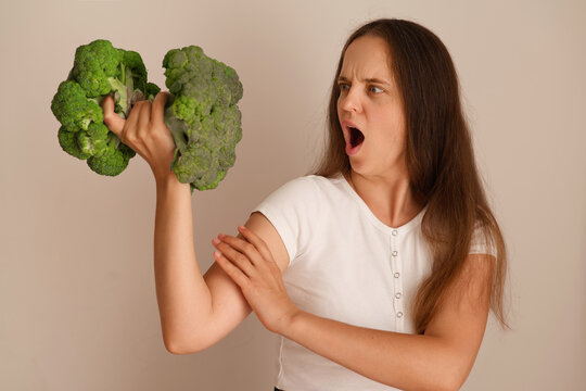 A woman lifts two heads of broccoli with a strong grip while making a facial expression that shows determination. She stands against a plain background