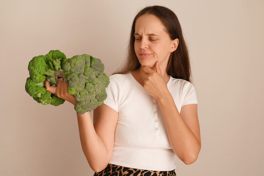 A woman stands indoors holding broccoli with a thoughtful expression. She considers the choice of healthy food while dressed in casual clothes