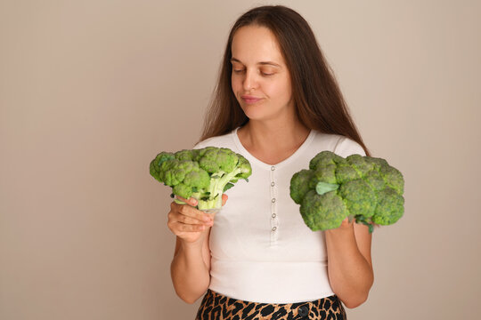 A woman stands in a neutral area holding one bunch of broccoli in each hand. She looks happy as she enjoys the healthy vegetables
