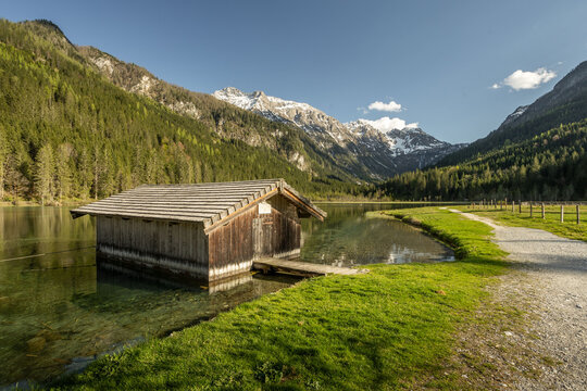 Wooden boathouse on the clear waters of Lake Jaegersee, surrounded by green forests and majestic snow-capped mountains under a bright blue sky, Kleinarl, Austria.