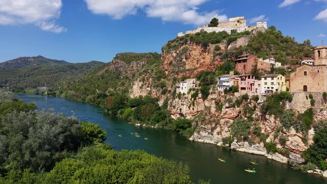 Aerial view of kayakers paddling the green Ebro River beneath the medieval Templar castle and cliffside village of Miravet, Tarragona, Catalonia, Spain