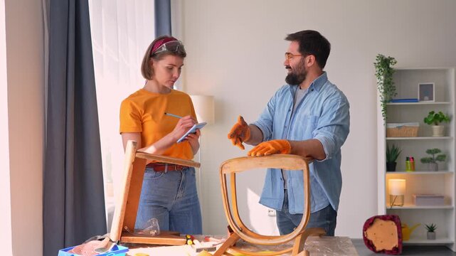 Happy young couple upcycling and restoring a vintage wooden chair. The man is sanding wood while the woman takes notes for their DIY project
