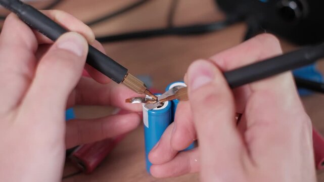 Close-up macro of hands spot welding nickel strips to blue lithium-ion battery terminals. The DIY assembly process highlights precision engineering in a workshop environment.