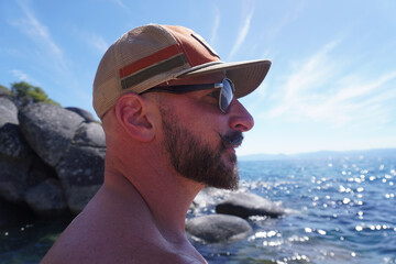 Side profile portrait of a bearded man wearing sunglasses and a trucker cap while standing beside a crystal-clear mountain lake. Bright summer weather, outdoor recreation, travel, relaxation, and natu © Christopher