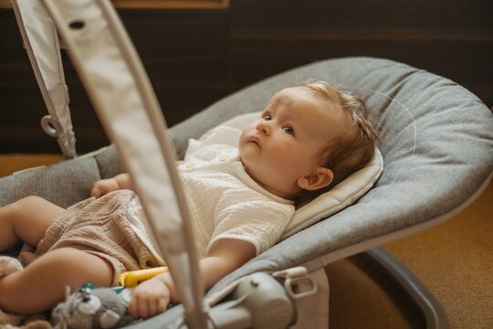 Cute baby lying in comfortable bouncer seat at home, looking up curiously. Cozy lifestyle, warm neutral colors.