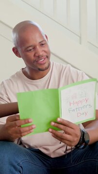 Vertical video: Presenting child giving green Father's Day card, father reading on couch, smiling
