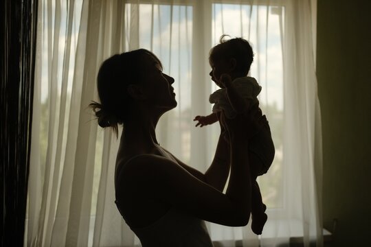 ilhouette of a mother lifting her baby up in front of a window, cozy home atmosphere.