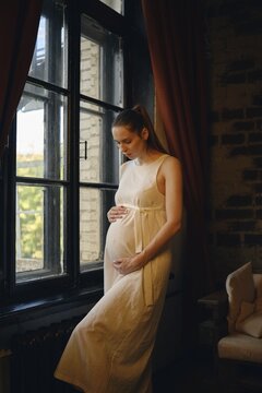 A woman in a light-colored dress stands near a sunny window, gently cradling her baby bump. She hugs her belly and talking to a baby