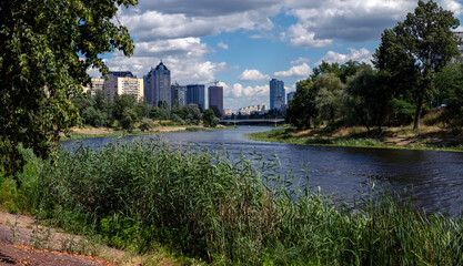 Fototapeta premium Cityscape showcasing modern apartment buildings reflecting urban development, coexisting with a wide winding river, lush green trees, and natural vegetation under a cloudy sky