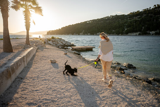 Woman walking black Labrador retriever along seaside path