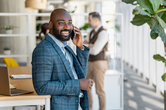 Smiling businessman on phone in modern office leaning on desk with laptop and plants
